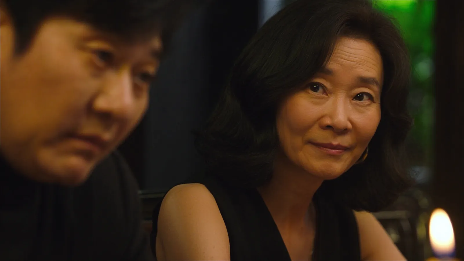 A woman in a black dress smiling quietly toward the camera at a candlelit table while a man beside her looks down