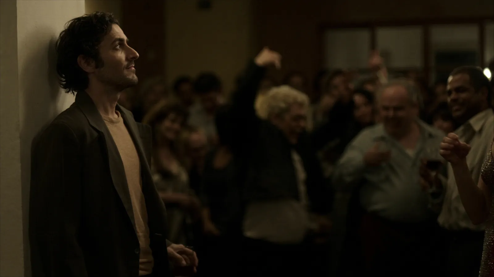 A man leaning against a wall at a crowded party, looking aside while guests laugh and cheer behind him