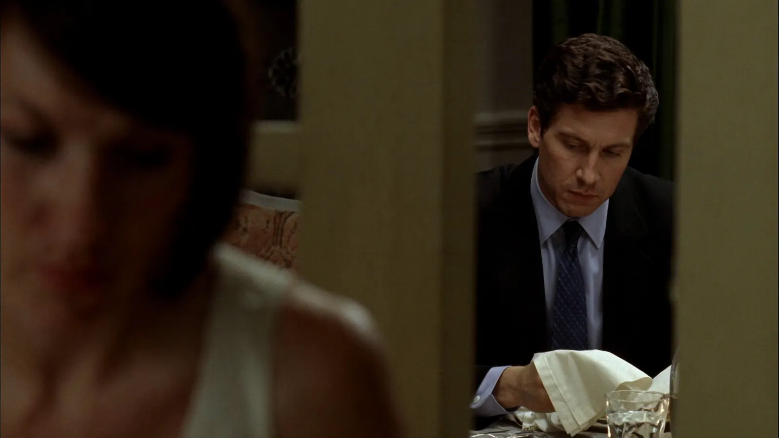 A man in a suit sitting composed at a dining table with a napkin in hand, while a woman looks down, out of focus, in the foreground