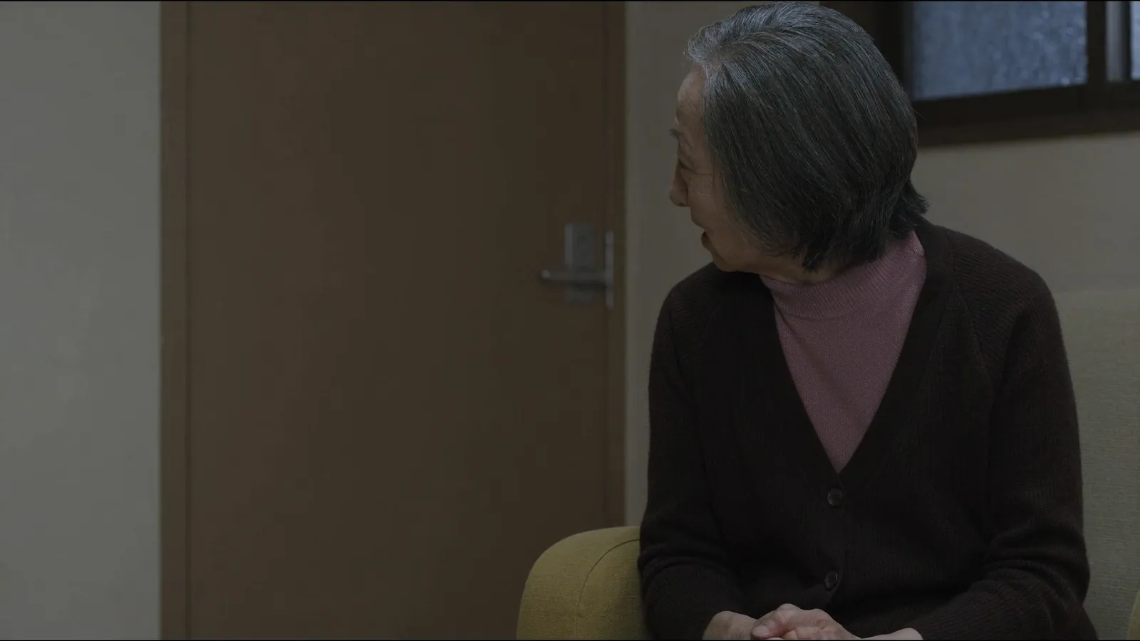 An elderly woman in a cardigan seated beside a door, turning to look at someone out of frame