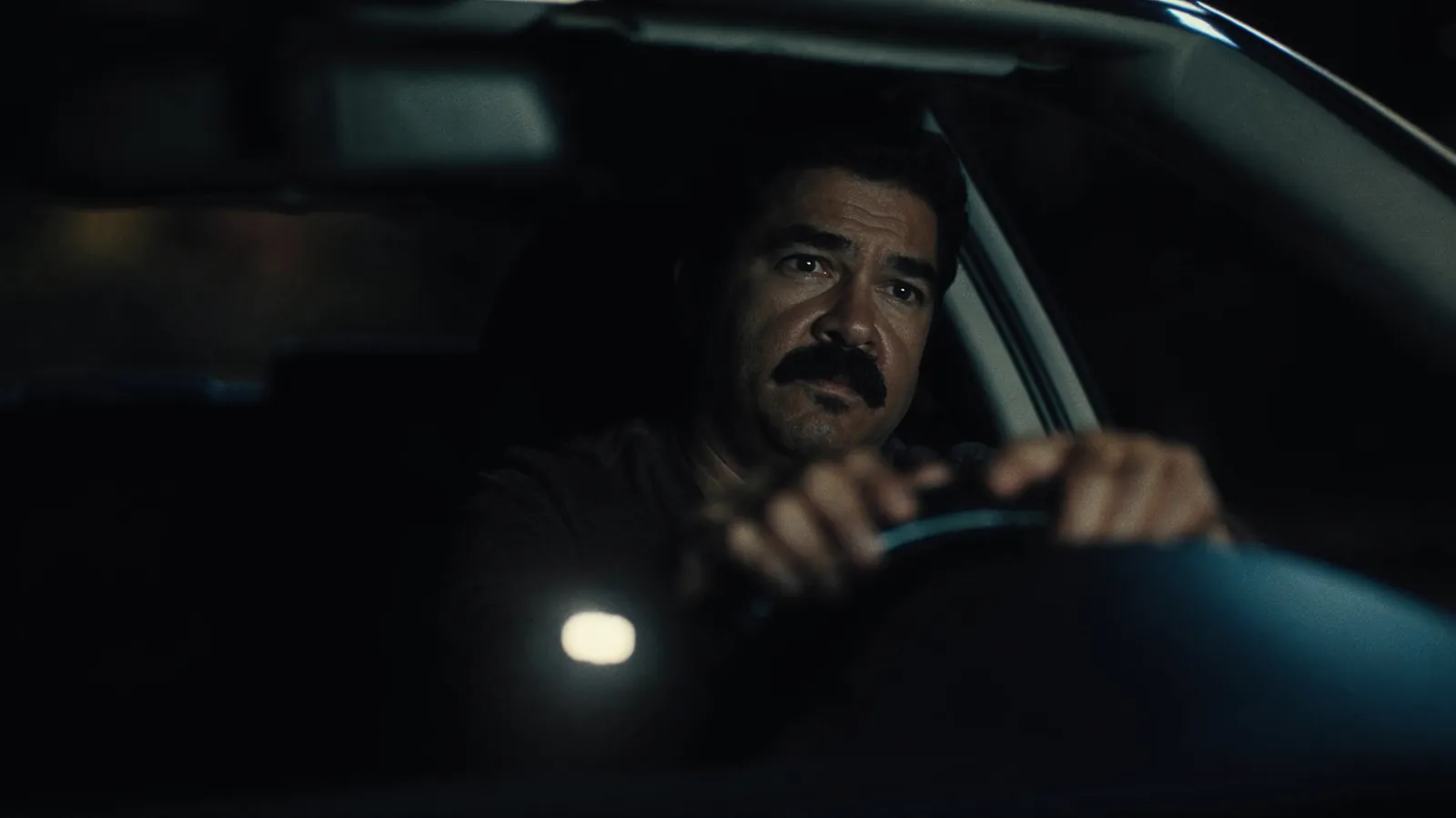 A mustached man gripping the steering wheel of a car at night, staring ahead