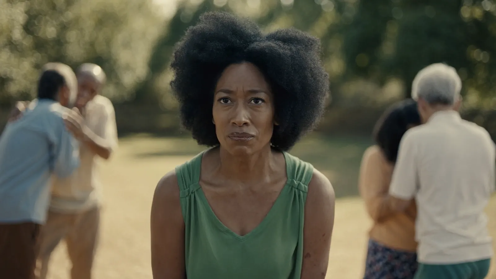 A woman in a green top standing alone with a hurt, tense look while two pairs of older people embrace and whisper behind her