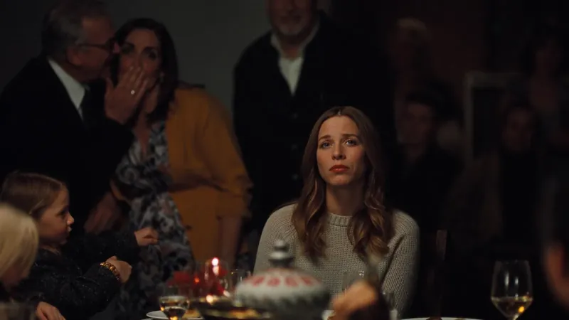 A woman sitting tense and alone at a family dinner table while relatives whisper behind her