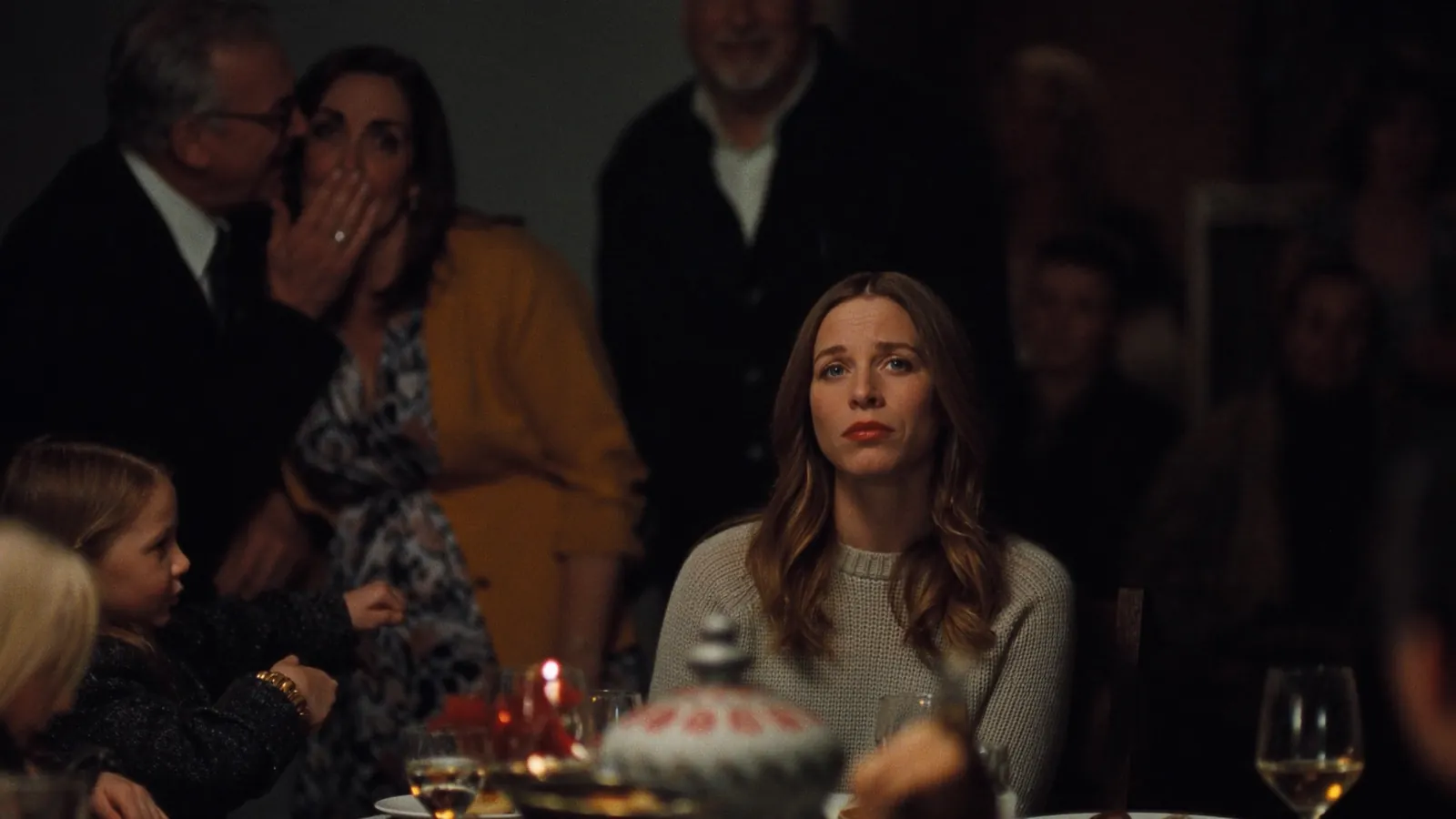 A woman sitting tense and alone at a family dinner table while relatives whisper behind her