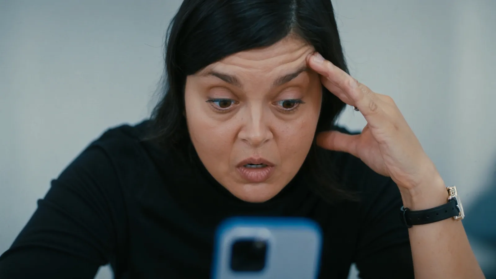 A woman with a hand pressed to her temple, wide-eyed and confused as she stares at her phone