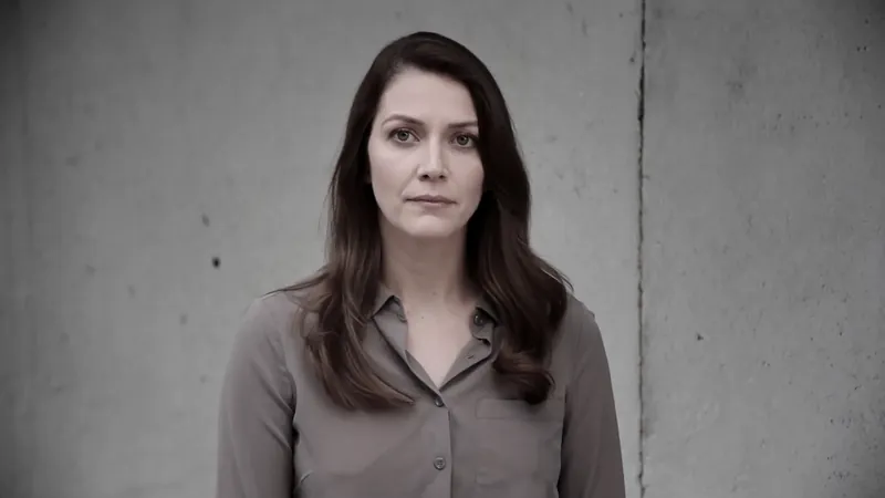 A woman standing against a bare concrete wall with a calm, expressionless face