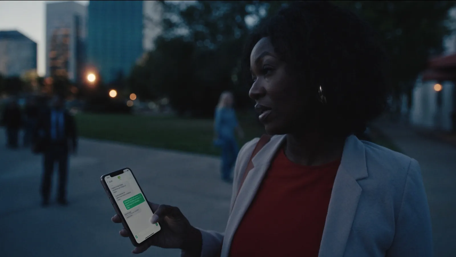 A woman in a blazer standing on a city path at dusk, holding a phone with a text conversation on screen