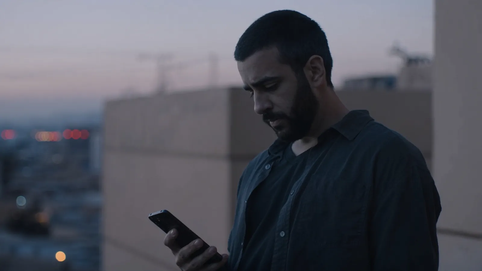 A bearded man standing on a rooftop at dusk, looking down at the phone in his hand