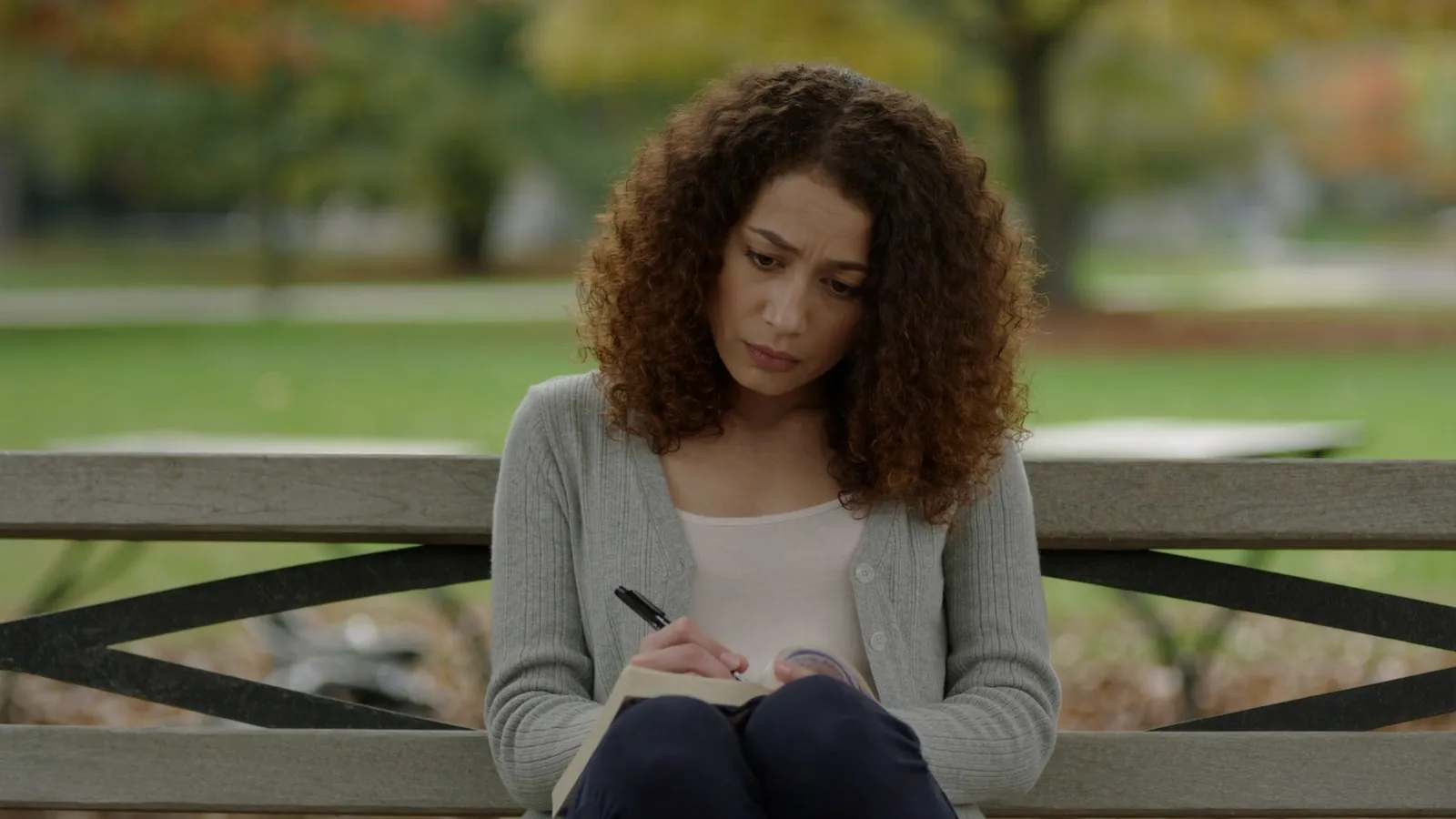 A young woman with curly hair sitting on a park bench, writing in a notebook with a troubled expression