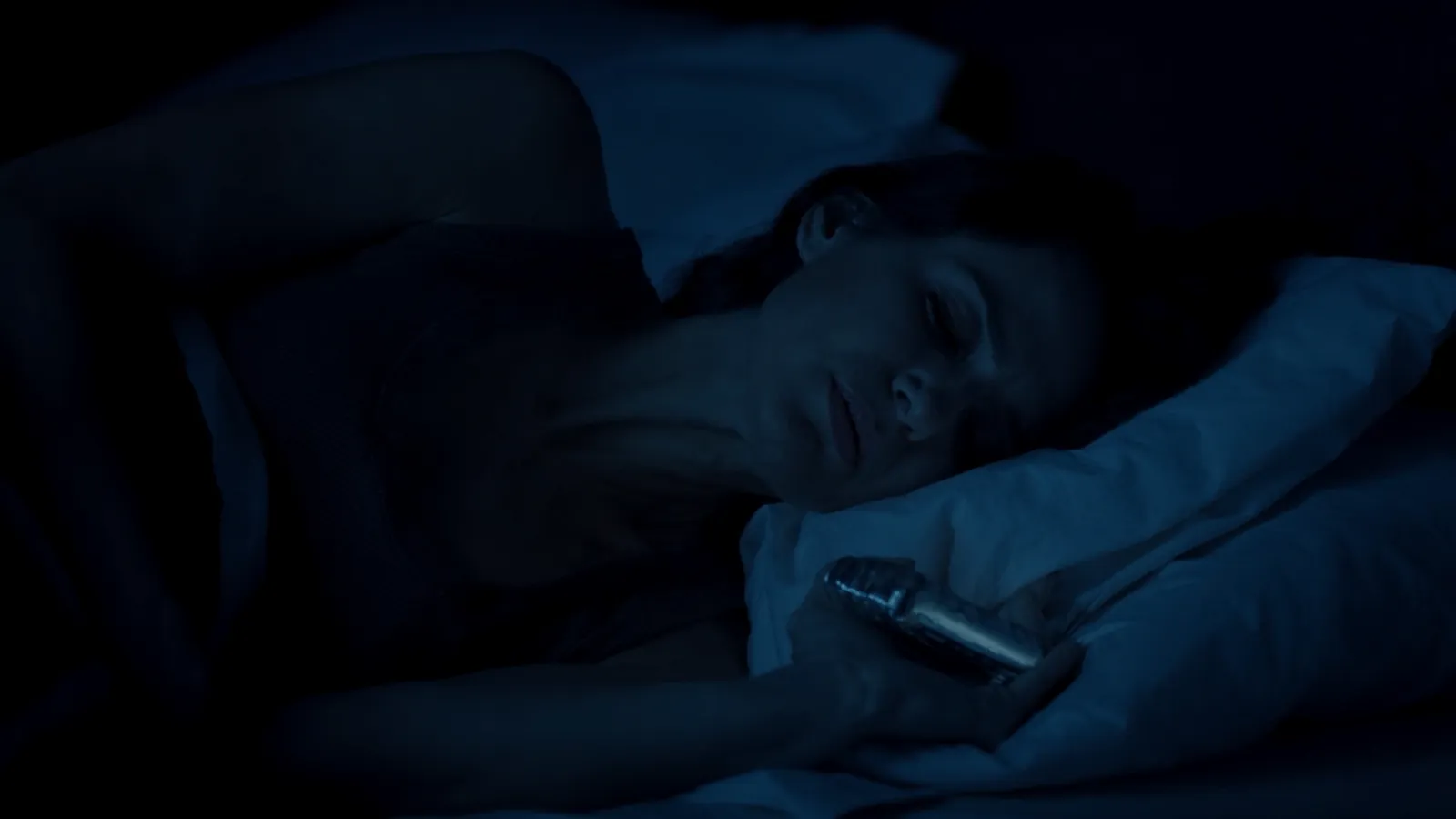 A woman lying awake in bed at night in a dark blue room, a phone resting on the pillow next to her hand