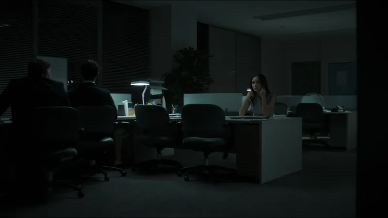 A woman on the phone at a desk in an otherwise dark open-plan office, two colleagues in silhouette nearby