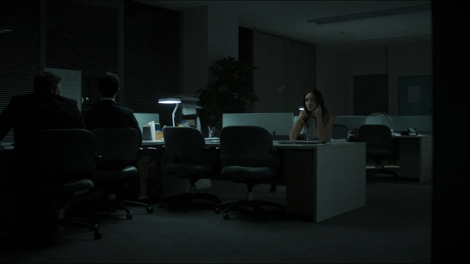 A woman on the phone at a desk in an otherwise dark open-plan office, two colleagues in silhouette nearby