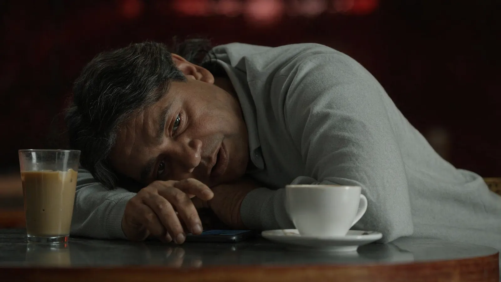 A man resting his head on his arm at a café table beside a coffee and a glass, a phone under his hand