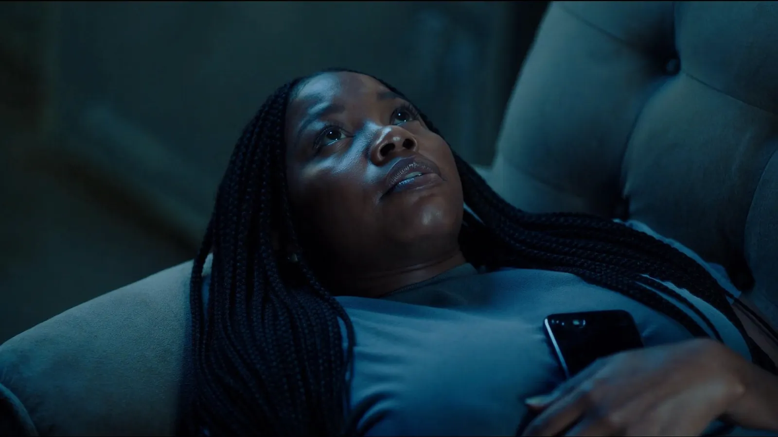 A woman lying on a sofa in a dark blue room, gazing upward with a phone resting on her chest