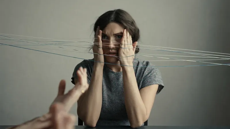 A woman covering her face with both hands, wide-eyed, as threads are pulled tight across the frame and hands reach toward her