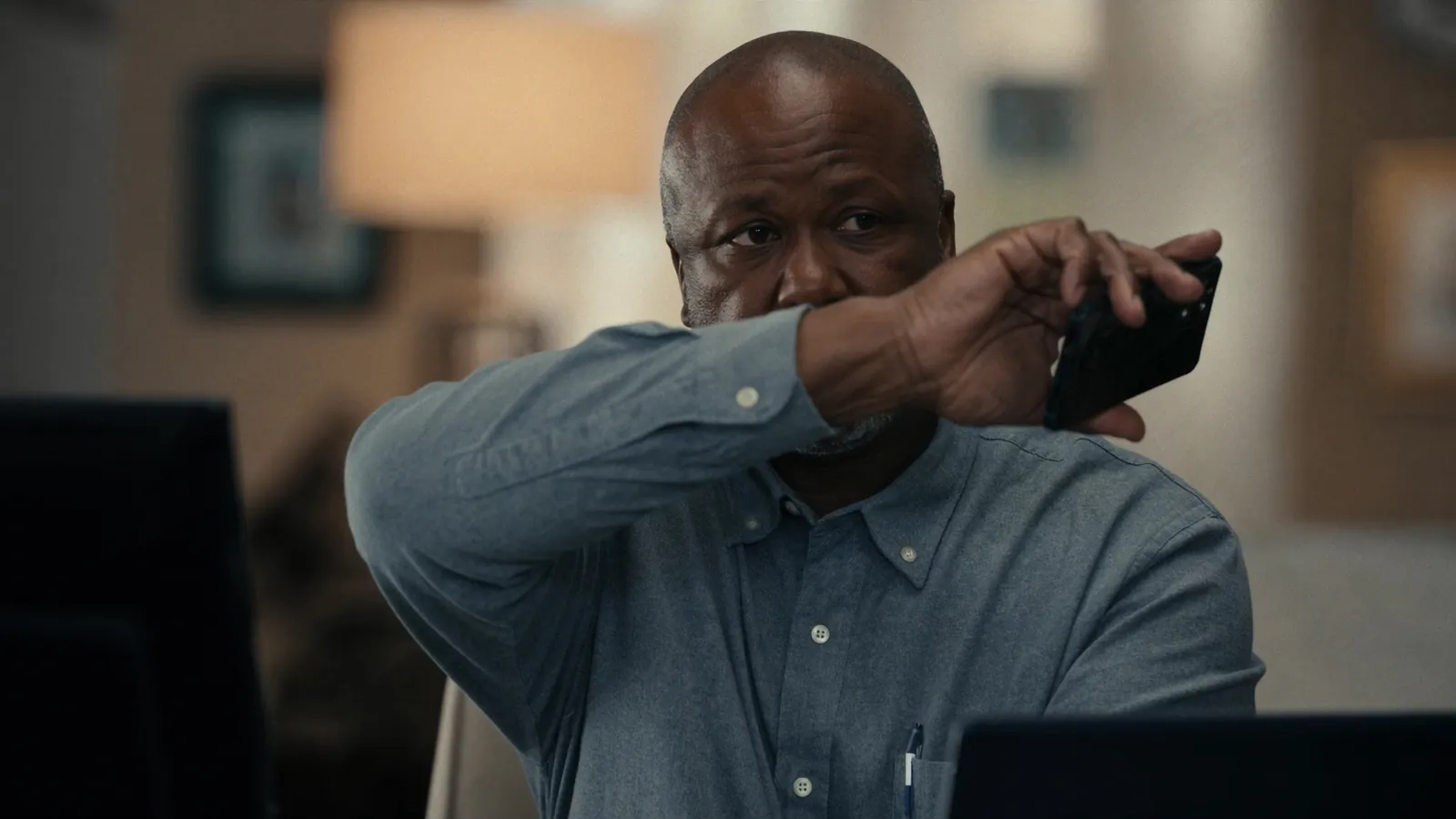 A man in a blue shirt wiping his face with one hand while holding a phone, seated at a desk in a dim room