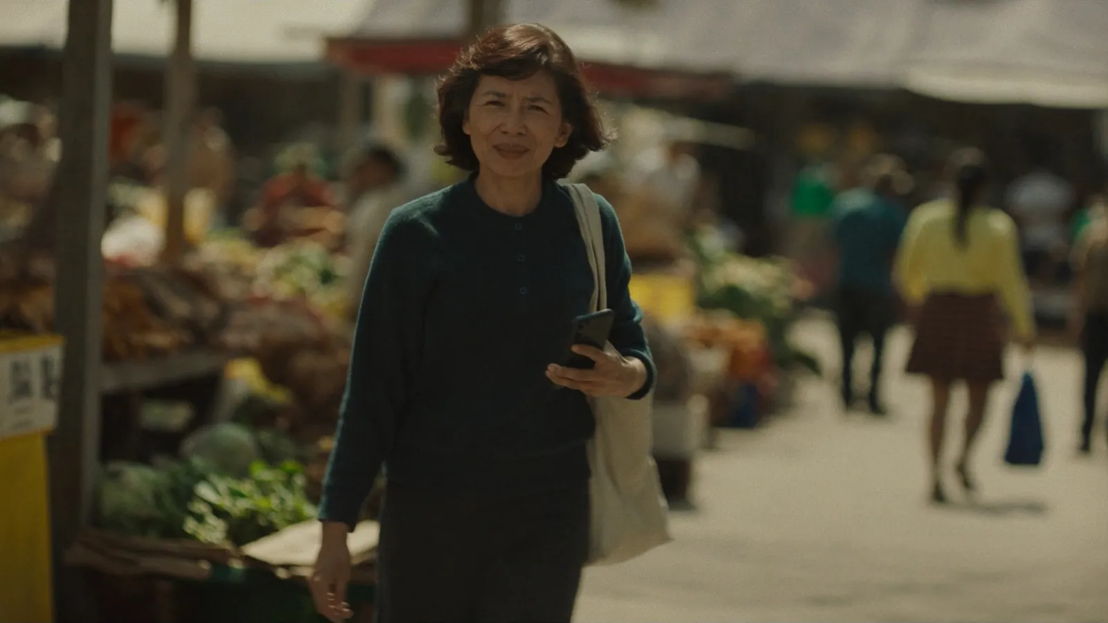 A woman with a tote bag walking through an outdoor market, phone in hand and a tense expression