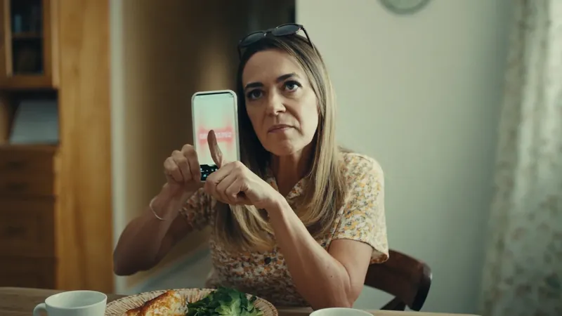 A woman at a dining table holding her phone up to the camera with a steady, skeptical stare