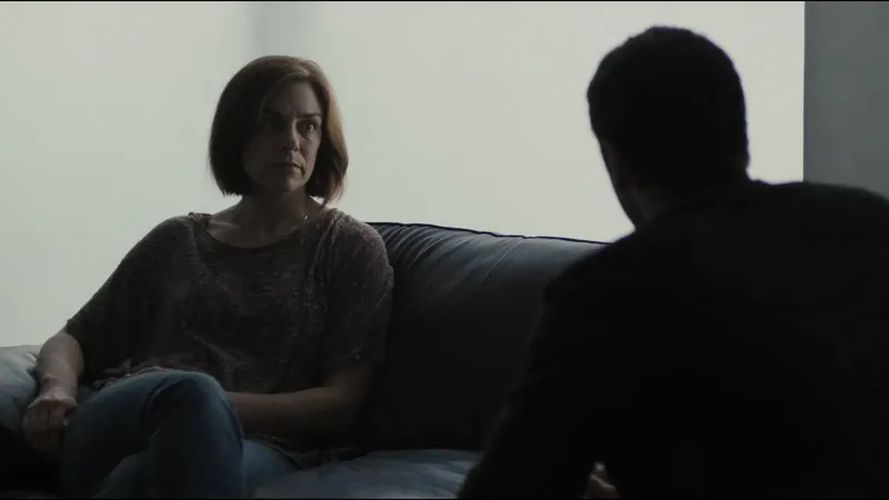 A woman sitting at one end of a sofa looking away, while a man sits in silhouette with his back to her