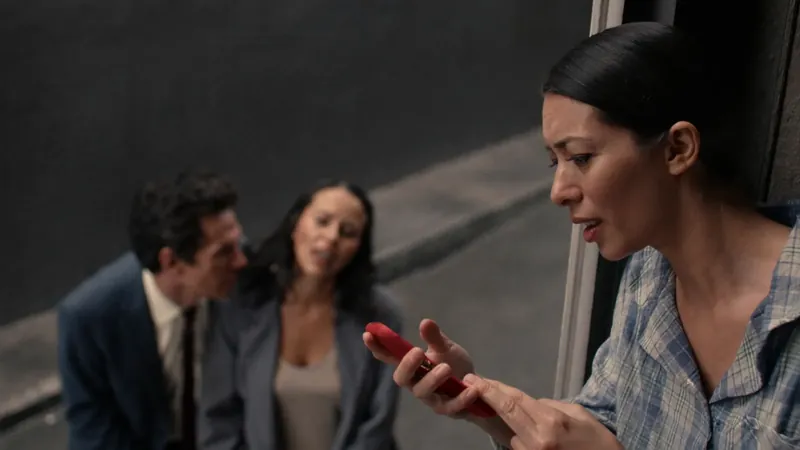 A woman in pajamas leaning out of a doorway, upset and speaking at her phone, while a man and a woman whisper close together behind her