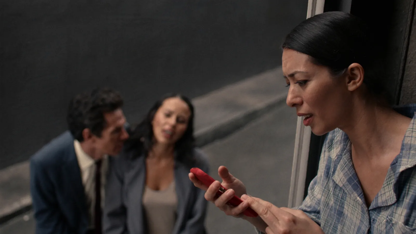 A woman in pajamas leaning out of a doorway, upset and speaking at her phone, while a man and a woman whisper close together behind her