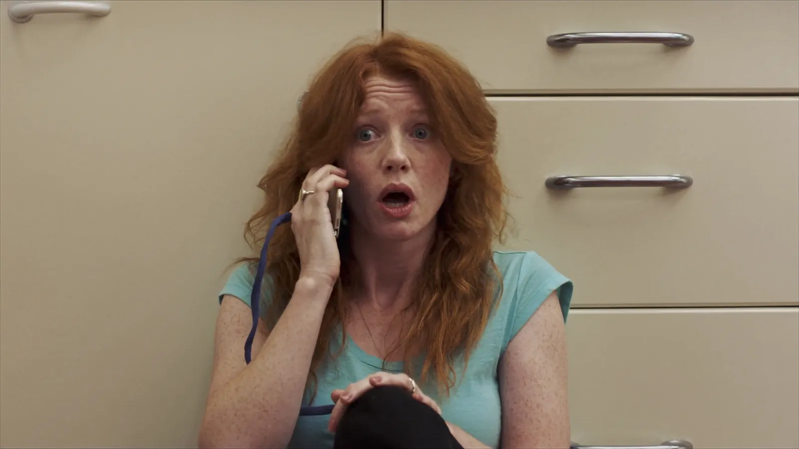 A red-haired woman sitting on a kitchen floor against the cabinets, phone to her ear and mouth open in shock