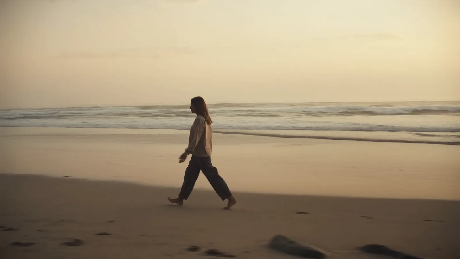 A woman walking barefoot along an empty beach at sunrise with the sea behind her