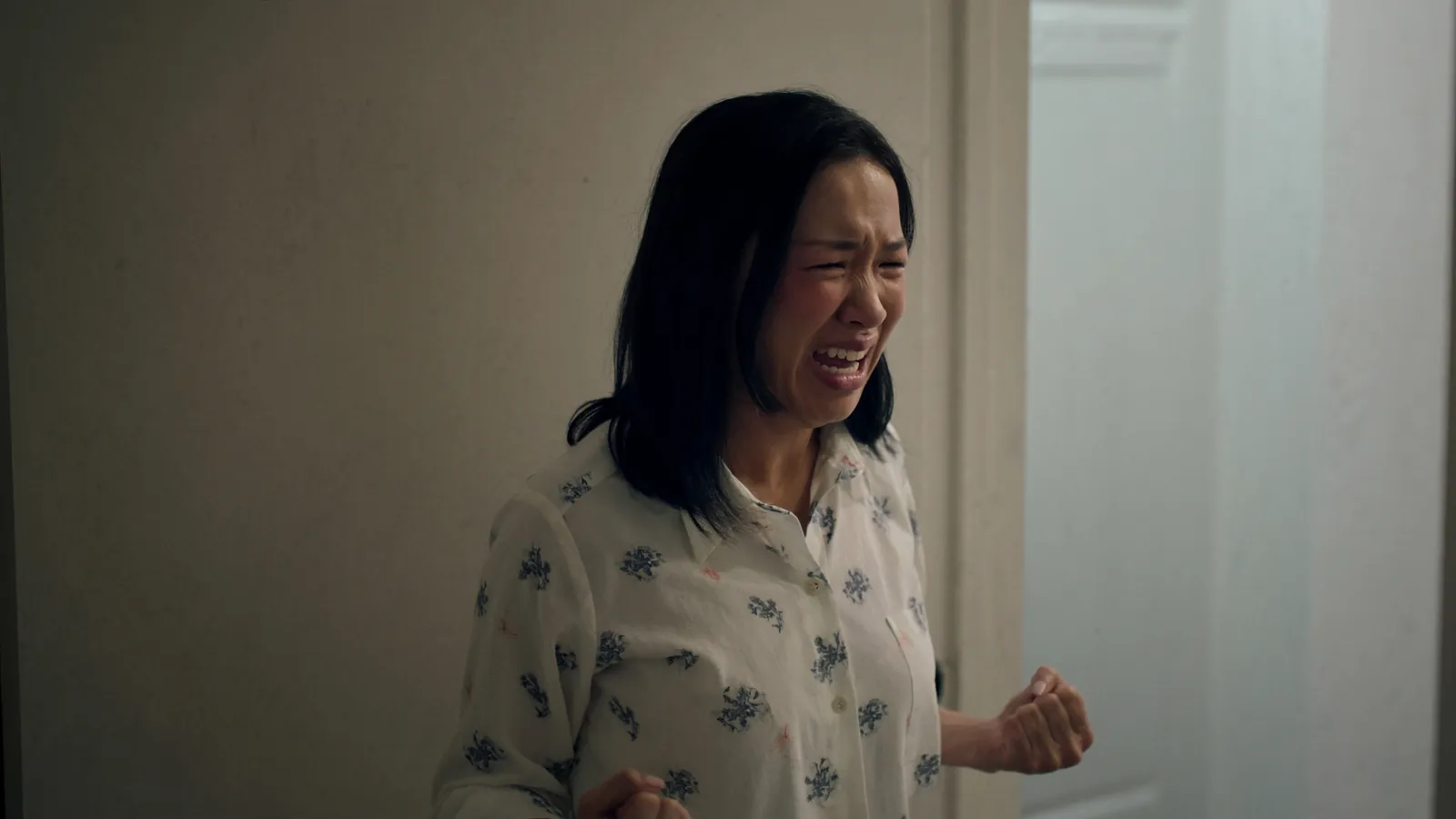 A woman in pajamas standing in a doorway, her face crumpled in tears and one fist clenched