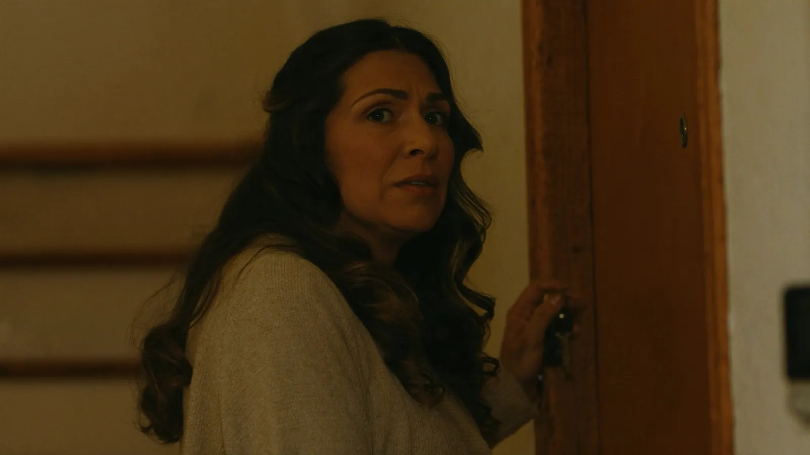 A woman with long wavy hair glancing warily over her shoulder, one hand on a wooden door and keys in her fingers