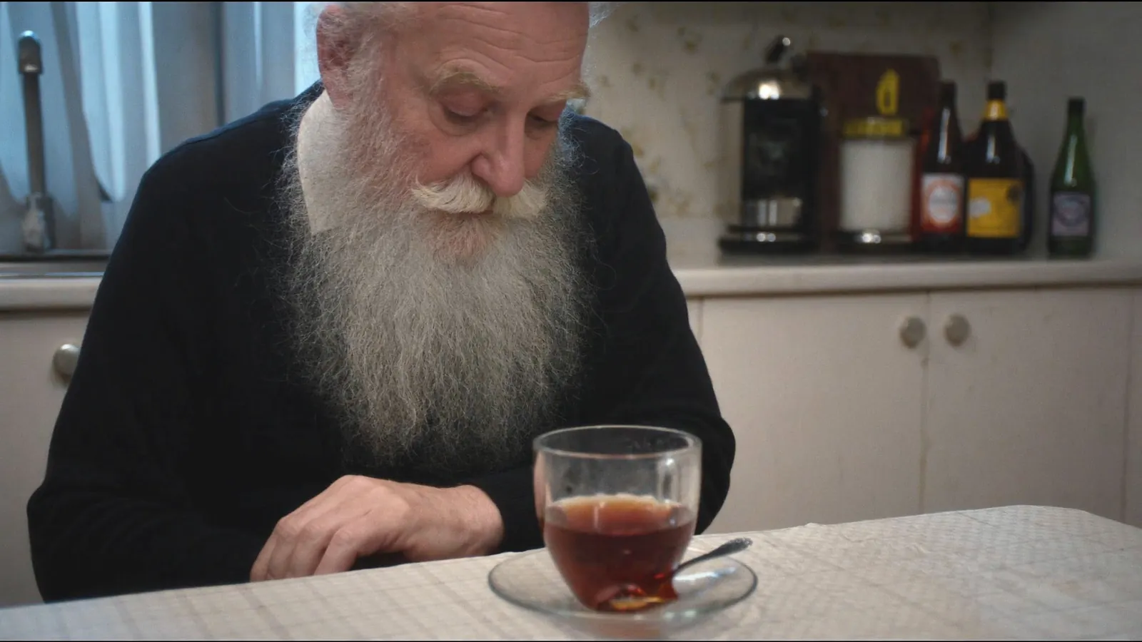 An older man with a long white beard looking down at a glass of tea on a kitchen table