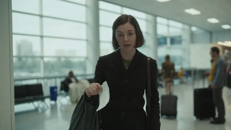 A woman walking through an airport terminal with a bag on her shoulder and a sheet of paper in her hand, looking tired and distant
