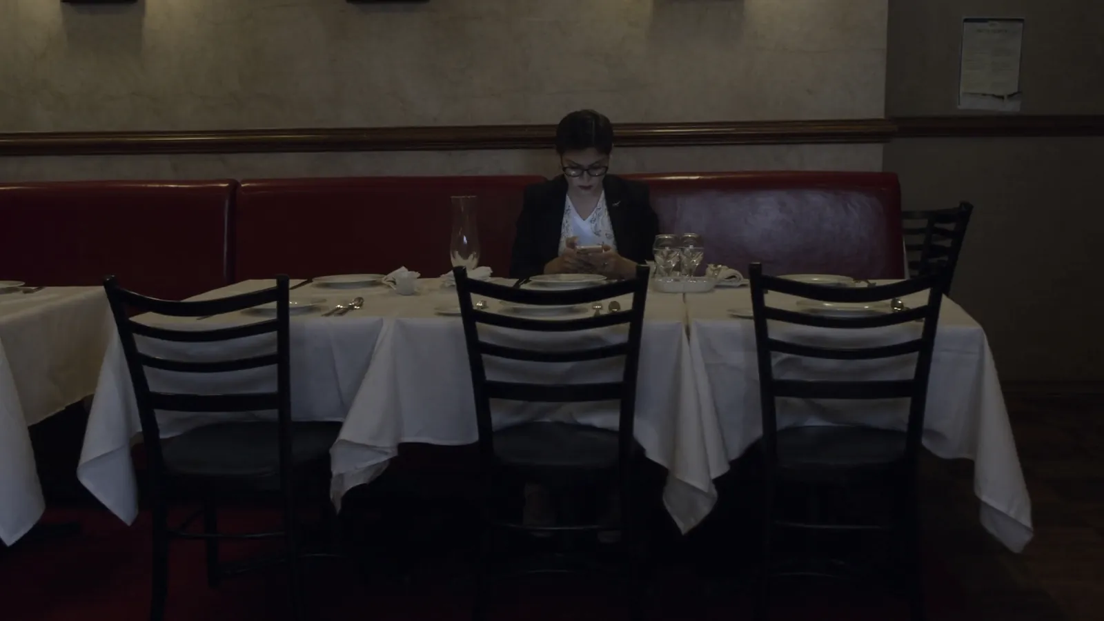 A woman sitting alone at a restaurant table set for several people, looking at her phone among empty chairs