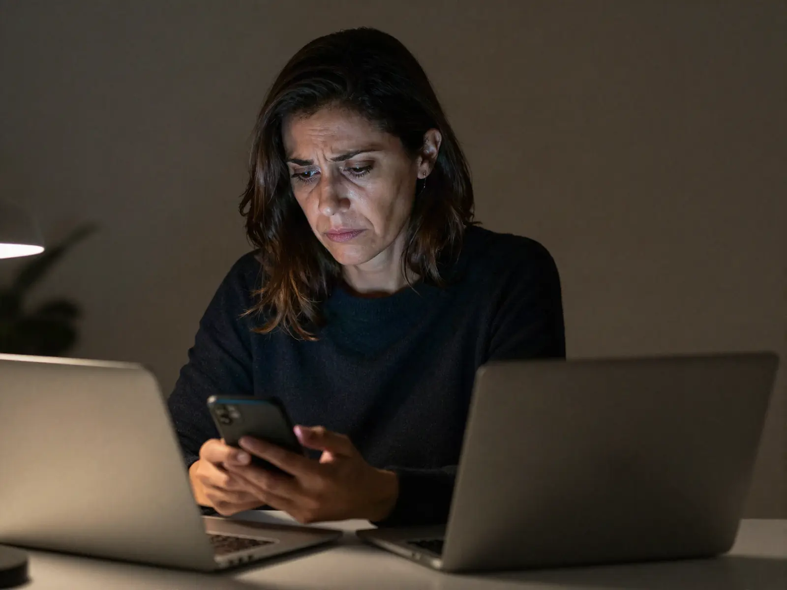 A woman at a laptop, rereading messages on her phone late at night