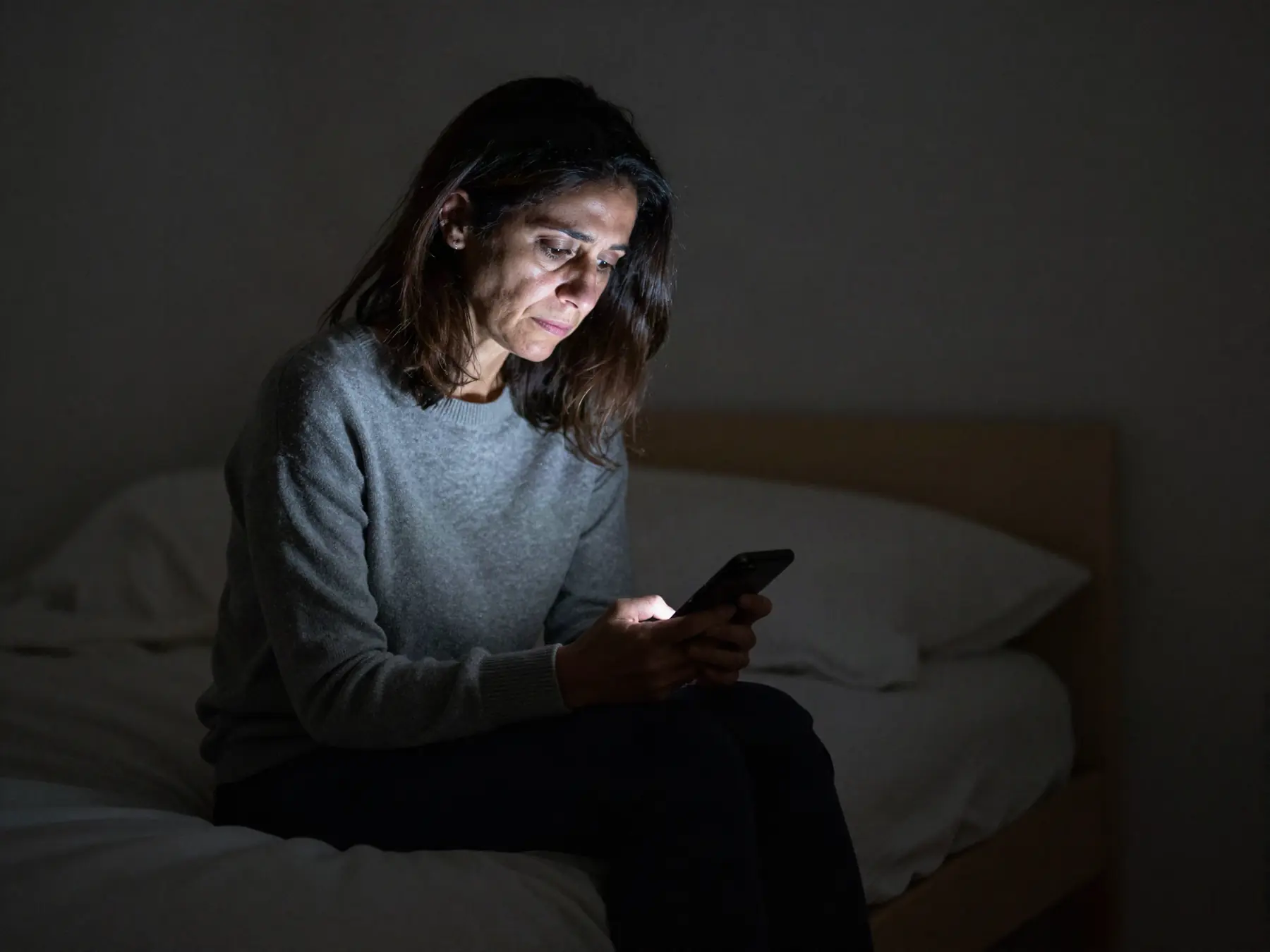 A woman sitting on her bed at night, her face lit by her phone screen