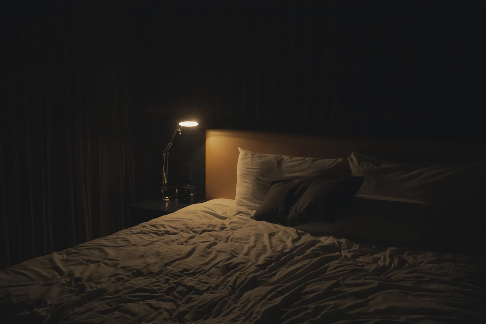 A dim bedroom at night with a small lamp and rumpled sheets