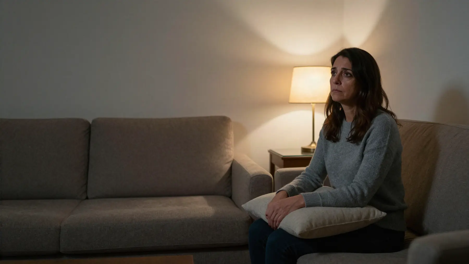 A woman sitting at one end of a living-room sofa with a cushion on her lap, looking into the distance