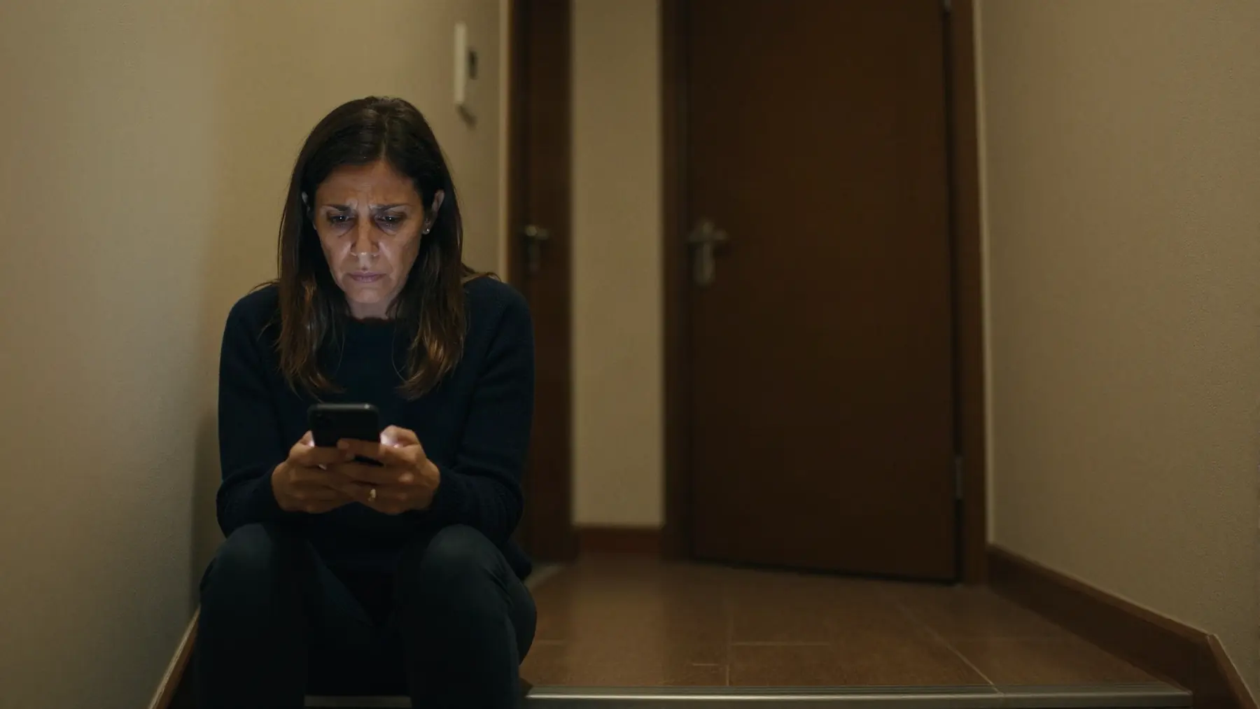 A woman sitting on a staircase landing, looking at her phone with a tired expression